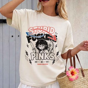 May include: Cream-colored t-shirt with a graphic design. The design features the words "STUPID FUCKING PINKS" with an illustration of a character holding a knife and bow and arrow. The shirt also has lightning bolt graphics.