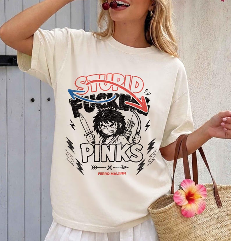 May include: Cream-colored t-shirt with a graphic design. The design features the words "STUPID FUCKING PINKS" with an illustration of a character holding a knife and bow and arrow. The shirt also has lightning bolt graphics.