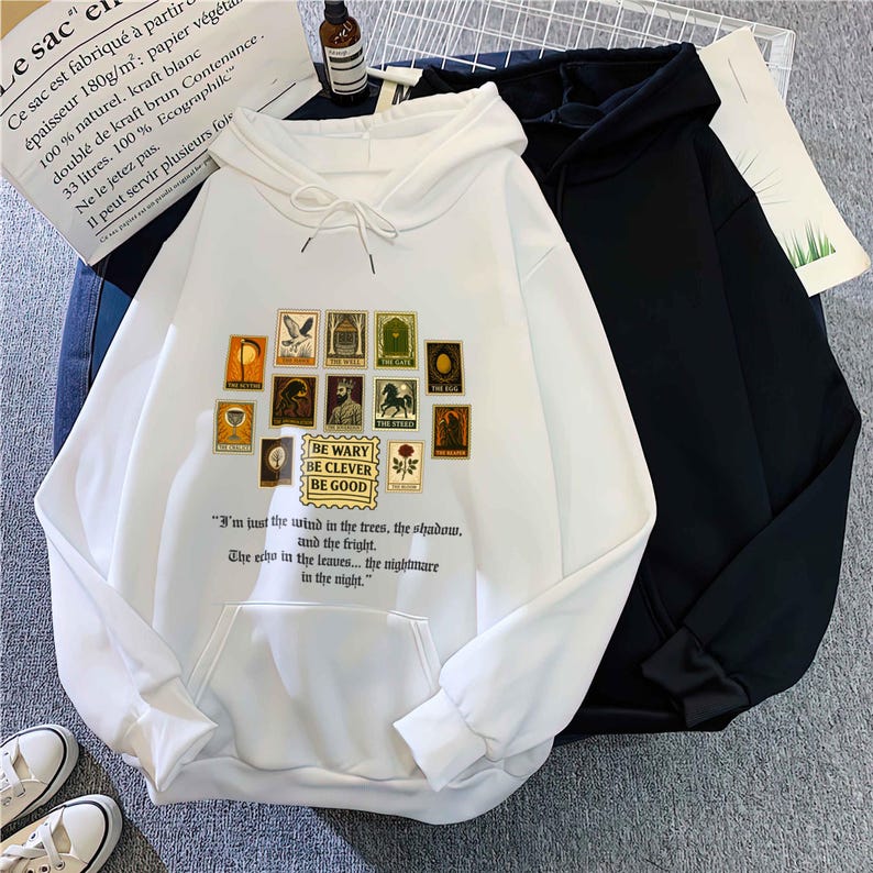 May include: White hoodie with a front pocket and drawstring hood. The front features a collection of vintage-style postage stamp illustrations and the text "BE WARY BE CLEVER BE GOOD". A black hoodie is next to it.