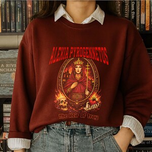 Op de afbeelding: Bordeaux sweatshirt met de tekst "ALEXIA PYROGENNETOS" in het rood. De afbeelding toont een gekroonde vrouw met een kruis, omringd door vlammen en een schedel. De tekst "The Heir of Troy" staat eronder.