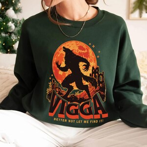 May include: Dark green sweatshirt featuring a graphic of a werewolf against a red moon and a castle silhouette. The word "Vigga" is displayed in orange and white, with the text "Better not let me find it!" below.