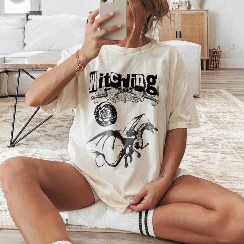 May include: A cream-coloured t-shirt with the word "Witching" in a bold, black font. Below the text is a black and white illustration of a dragon with outstretched wings. The shirt also features a rose and sword design.