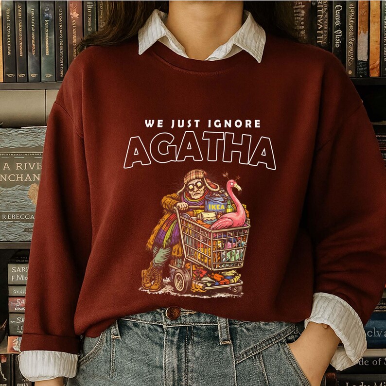 May include: Burgundy sweatshirt with the text "WE JUST IGNORE AGATHA" in white. The graphic depicts a person pushing a shopping cart filled with items, including a pink flamingo. The person is wearing a hat and glasses.