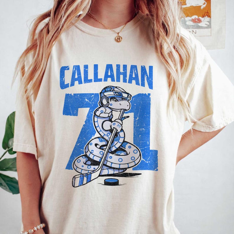May include: Cream-colored t-shirt featuring the name "CALLAHAN" in blue block letters above the number "71". A cartoon snake wearing a hockey helmet and holding a hockey stick is in the center. The design has a distressed, vintage look.
