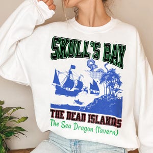 May include: White sweatshirt with a blue graphic featuring a pirate ship, palm trees, and the text "SKULL'S BAY" in green and black. Below, the text "THE DEAD ISLANDS" is in red and black, with "The Sea Dragon (Tavern)" in green.
