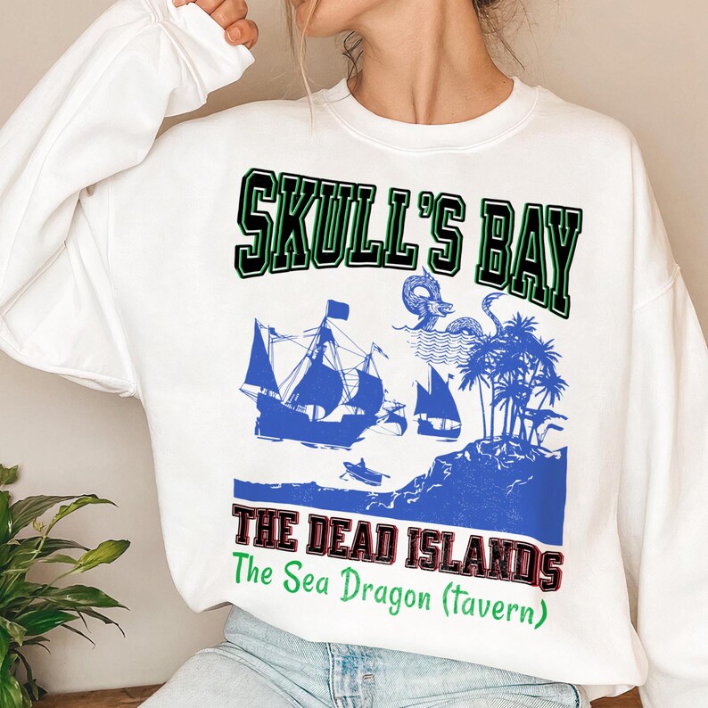 May include: White sweatshirt with a blue graphic featuring a pirate ship, palm trees, and the text "SKULL'S BAY" in green and black. Below, the text "THE DEAD ISLANDS" is in red and black, with "The Sea Dragon (Tavern)" in green.