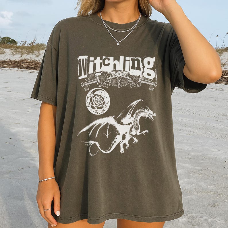 May include: Dark olive green t-shirt featuring the word "Witchling" in a distressed font, a white dragon graphic, a rose, and crossed swords. The shirt has short sleeves and a loose fit.