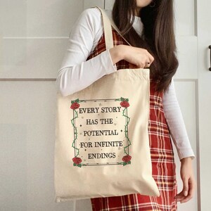 May include: A beige tote bag with a black and white graphic that reads "Every story has the potential for infinite endings". The graphic is surrounded by a black border and red roses with green vines.