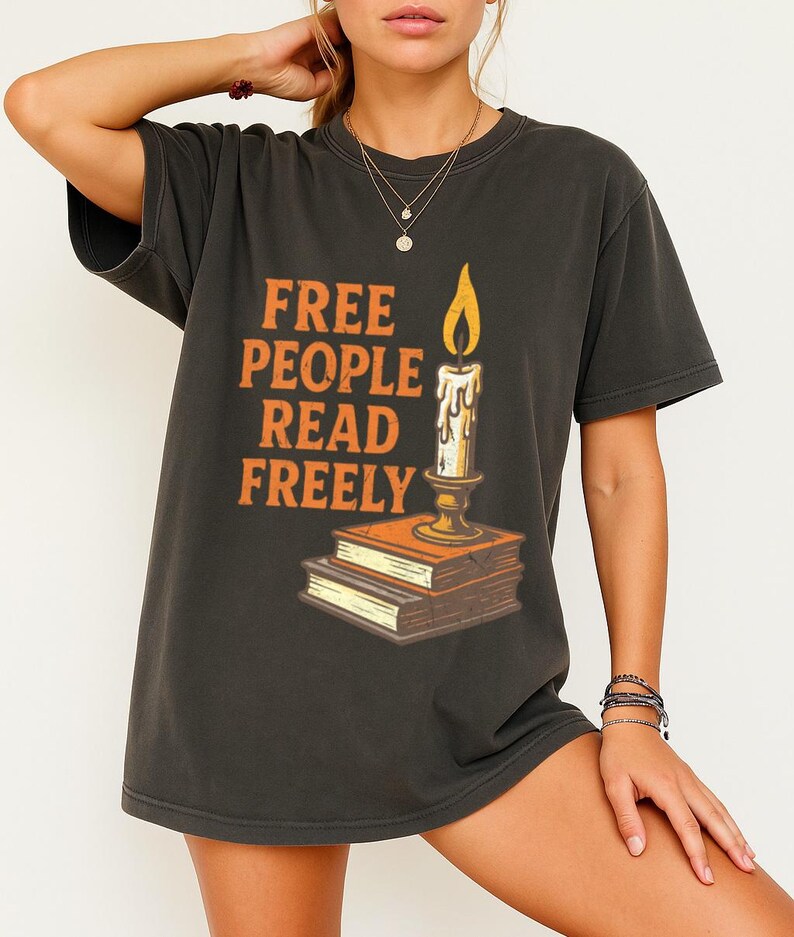 Może przedstawiać: Ciemnoszara koszulka z pomarańczowym napisem "FREE PEOPLE READ FREELY". Świeca w świeczniku stoi na stosie książek. Płomień świecy jest ż&oacute;łty.