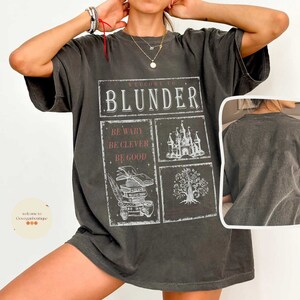 Op de afbeelding: Een oversized donkergrijs T-shirt met een grafische print die "Welkom bij Blunder" zegt en een kasteel, een boom en een stapel boeken toont. De tekst onder het kasteel zegt "Wees voorzichtig, wees slim, wees goed."