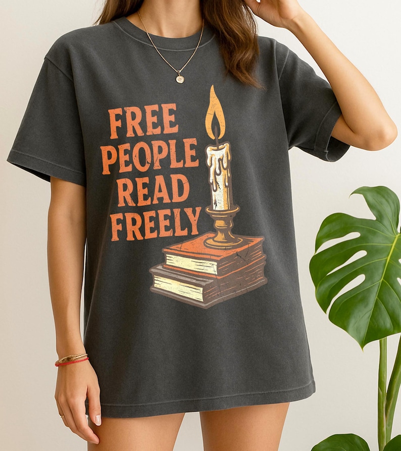 Może przedstawiać: Ciemnoszara koszulka z pomarańczowym napisem "FREE PEOPLE READ FREELY". Grafika przedstawia świecę na dw&oacute;ch książkach. Koszulka ma okrągły dekolt i kr&oacute;tkie rękawy.