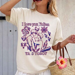 May include: Cream-colored t-shirt with a graphic design featuring two figures embracing, surrounded by illustrations of a snake, hearts, a spiderweb, and other elements. The text "I love you Yulian" and "YA S TOBOY" are also included.