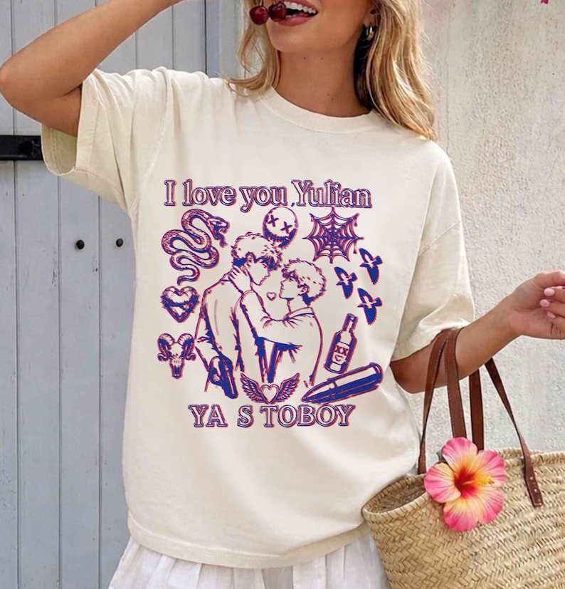 May include: Cream-colored t-shirt with a graphic design featuring two figures embracing, surrounded by illustrations of a snake, hearts, a spiderweb, and other elements. The text "I love you Yulian" and "YA S TOBOY" are also included.