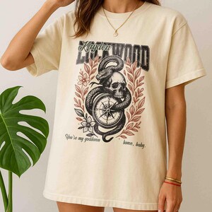 Puede incluir: Camiseta de color crema con un dise&ntilde;o gr&aacute;fico que presenta una calavera, una serpiente y una br&uacute;jula. El dise&ntilde;o incluye el texto "Kayden Lillywood" y la frase "You're my goddamn home, baby." La camiseta tiene mangas cortas.