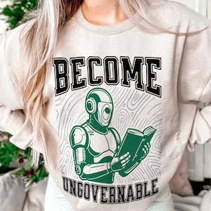 May include: Beige sweatshirt featuring the text "BECOME UNGOVERNABLE" in black. A green robot reading a book is in the centre. The background has a blue, wavy design.
