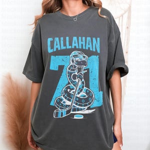 May include: Dark gray t-shirt featuring the name "CALLAHAN" in light blue block letters above the number "71". The number "71" has a light blue snake graphic wearing a hockey helmet and holding a hockey stick. The shirt is a casual, oversized fit.