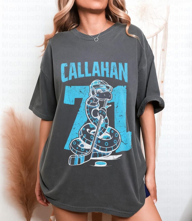 May include: Dark gray t-shirt featuring the name "CALLAHAN" in light blue block letters above the number "71". The number "71" has a light blue snake graphic wearing a hockey helmet and holding a hockey stick. The shirt is a casual, oversized fit.