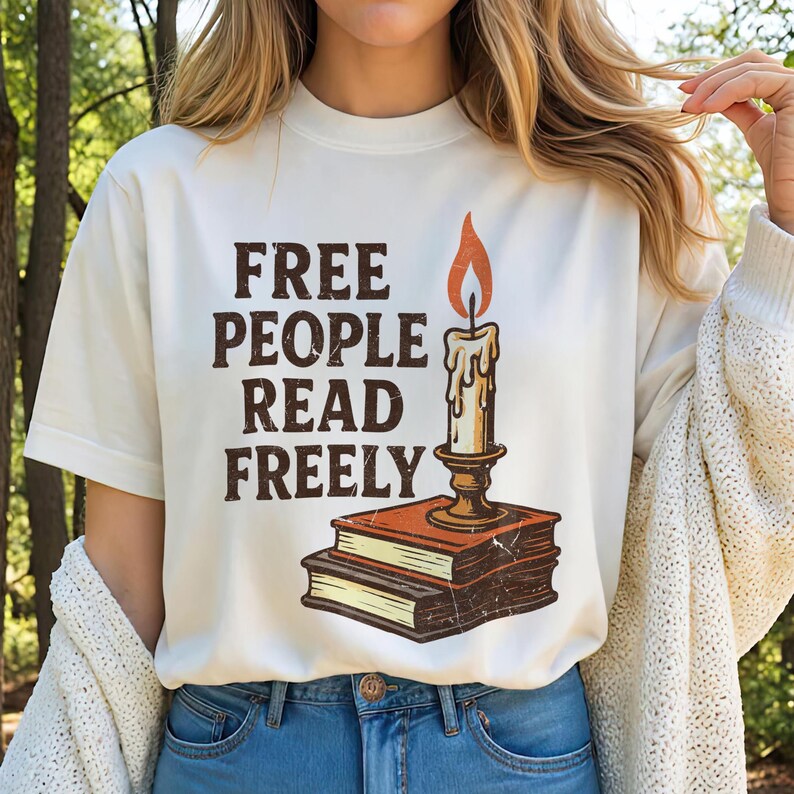Może przedstawiać: Kremowa koszulka z napisem "FREE PEOPLE READ FREELY" w brązowych literach. Świeca z płomieniem i kapiącym woskiem jest nadrukowana nad dwoma książkami. Koszulka ma swobodny styl.