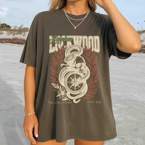 Puede incluir: Camiseta verde oliva oscuro con un dise&ntilde;o gr&aacute;fico. El dise&ntilde;o incluye una calavera, una serpiente, una br&uacute;jula y elementos florales en blanco, rojo y crema, con el texto "Kayden Lockwood". La camiseta tambi&eacute;n tiene el texto "You're my goddamn home, baby."