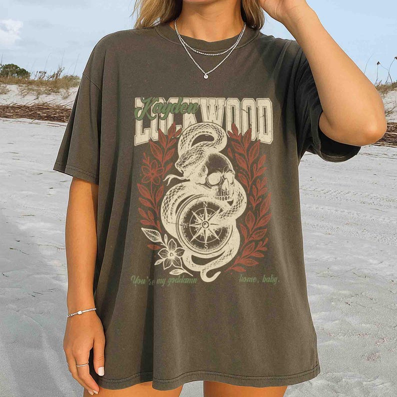 Puede incluir: Camiseta verde oliva oscuro con un dise&ntilde;o gr&aacute;fico. El dise&ntilde;o incluye una calavera, una serpiente, una br&uacute;jula y elementos florales en blanco, rojo y crema, con el texto "Kayden Lockwood". La camiseta tambi&eacute;n tiene el texto "You're my goddamn home, baby."