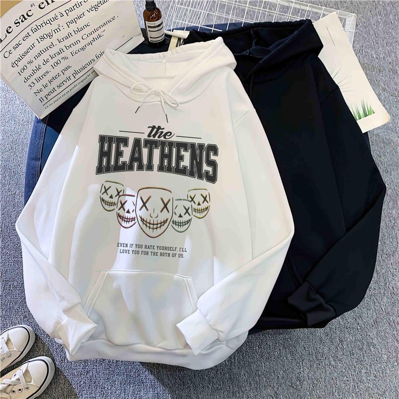 May include: A white hoodie featuring the text "The Heathens" and a graphic of three smiling masks. The hoodie has a front pocket and a drawstring hood. A black hoodie is next to it. The text "Even if you hate yourself, I'll love you for the both of us" is below the masks.