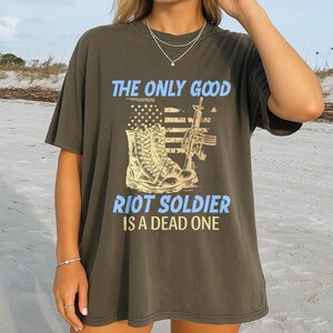 May include: Dark olive green t-shirt with the text "THE ONLY GOOD RIOT SOLDIER IS A DEAD ONE" in light blue. The graphic includes a combat boot, an assault rifle, and an American flag design.