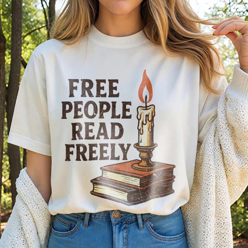 Może przedstawiać: Kremowy t-shirt z napisem "FREE PEOPLE READ FREELY" w brązowych literach. Na koszulce znajduje się grafika świecy na dw&oacute;ch książkach. Świeca ma pomarańczowy płomień.