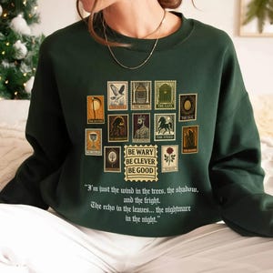 May include: Dark green sweatshirt featuring a collection of postage stamp-style graphics with various illustrations and text. The text "BE WARY BE CLEVER BE GOOD" is below the stamps. The sweatshirt also has a quote printed on it.