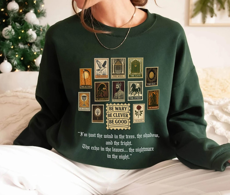 May include: Dark green sweatshirt featuring a collection of postage stamp-style graphics with various illustrations and text. The text "BE WARY BE CLEVER BE GOOD" is below the stamps. The sweatshirt also has a quote printed on it.