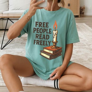 Może przedstawiać: Turkusowa koszulka z napisem "FREE PEOPLE READ FREELY" w brązowych literach. Grafika przedstawia świecę na dw&oacute;ch książkach. Świeca ma pomarańczowy płomień. Koszulka ma luźny, obszerny kr&oacute;j.
