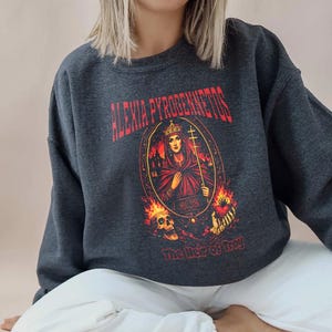 Op de afbeelding: Donkergrijze sweatshirt met een grafisch ontwerp. Het ontwerp bevat de tekst "ALEXIA PYROGENNETOS" in het rood en een ovaal portret van een gekroonde vrouw die een kruis vasthoudt. De tekst "The Heir of Troy" staat eronder.