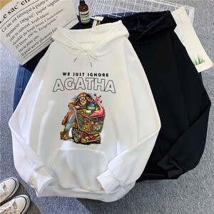 May include: White hooded sweatshirt with the text "WE JUST IGNORE AGATHA" above a colorful graphic of a person pushing a shopping cart. A black hooded sweatshirt is next to it. The white sweatshirt has a front pocket.