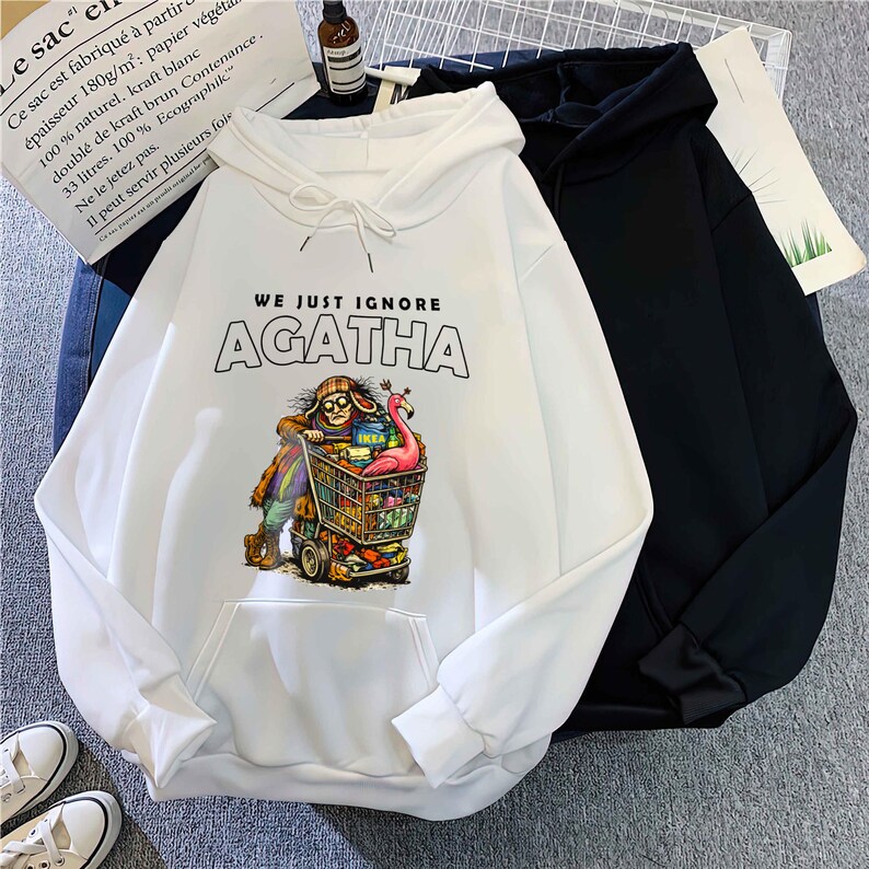 May include: White hooded sweatshirt with the text "WE JUST IGNORE AGATHA" above a colorful graphic of a person pushing a shopping cart. A black hooded sweatshirt is next to it. The white sweatshirt has a front pocket.