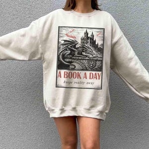 May include: Cream-colored sweatshirt featuring a black and white illustration of a dragon, castle, and the text "A BOOK A DAY keeps reality away." The graphic is in a rectangular frame. The sweatshirt has long sleeves.