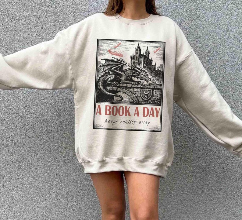 May include: Cream-colored sweatshirt featuring a black and white illustration of a dragon, castle, and the text "A BOOK A DAY keeps reality away." The graphic is in a rectangular frame. The sweatshirt has long sleeves.