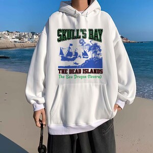 May include: White hooded sweatshirt with a front pocket and a drawstring. The graphic on the front features the text "SKULL'S BAY" above a blue illustration of ships, palm trees, and the text "THE DEAD ISLANDS The Sea Dragon (tavern)".