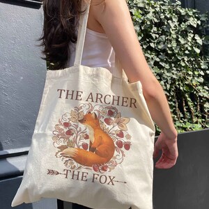 May include: A beige tote bag with a fox illustration and the text "The Archer The Fox".