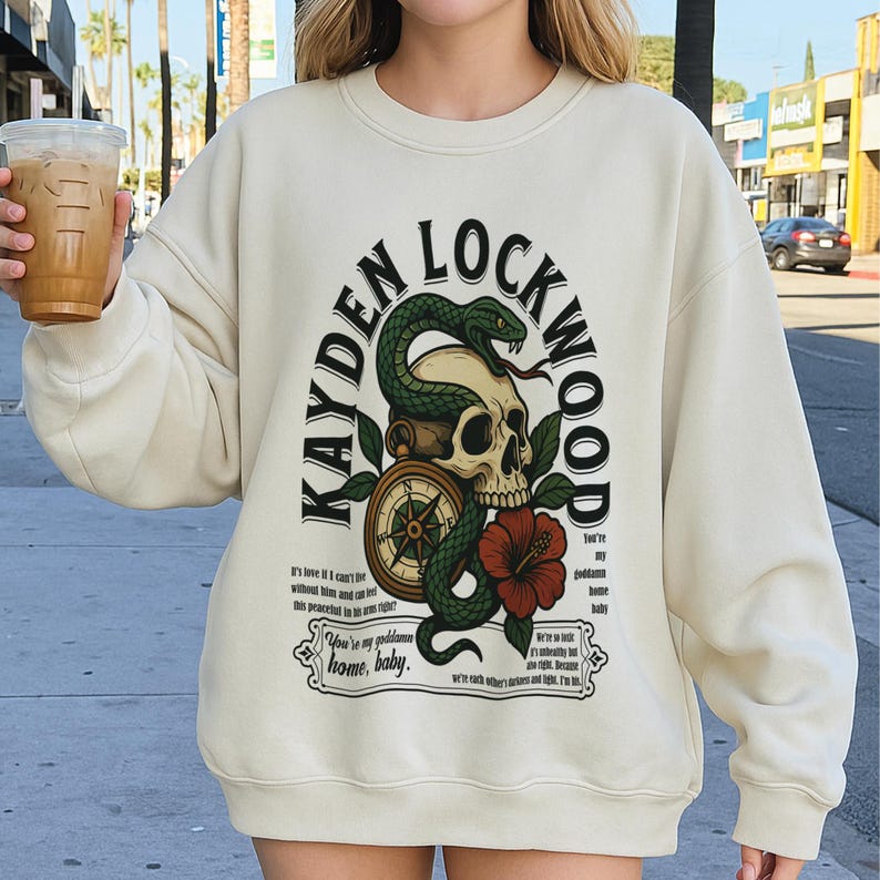 May include: Cream-colored sweatshirt with a graphic design featuring a skull, a green snake, a compass, and a red hibiscus flower. The text "KAYDEN LOCKWOOD" arches above the design. Additional text includes phrases like "You're my goddamn home, baby."