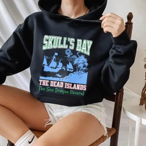 May include: Black hooded sweatshirt with a graphic design. The design features the text "Skull's Bay" above an illustration of a ship, palm trees, and the text "The Dead Islands The Sea Dragon (tavern)".