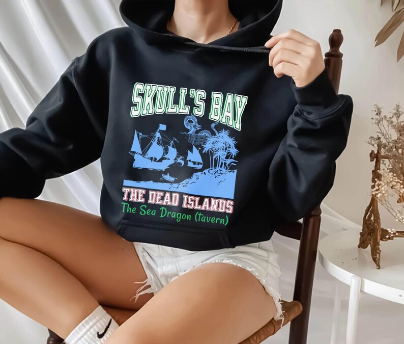 May include: Black hooded sweatshirt with a graphic design. The design features the text "Skull's Bay" above an illustration of a ship, palm trees, and the text "The Dead Islands The Sea Dragon (tavern)".