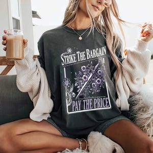 May include: A dark gray t-shirt with a graphic print of a sword and flowers. The text on the graphic reads "Strike the Bargain" and "Every choice has its consequences. Pay the Price."