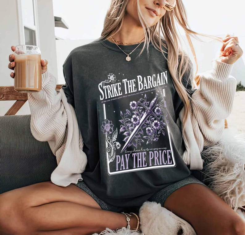 May include: A dark gray t-shirt with a graphic print of a sword and flowers. The text on the graphic reads "Strike the Bargain" and "Every choice has its consequences. Pay the Price."