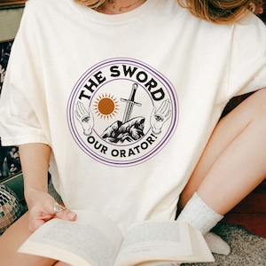 May include: Cream-colored t-shirt featuring a circular graphic with the text "THE SWORD OUR ORATOR!" in a purple and white design. The graphic includes a sword, sun, and hands with eyes.
