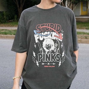 May include: Dark gray t-shirt with a graphic design. The design features the words "STUPID FUCKING PINKS" in red and white, with a cartoon character holding knives and arrows. The shirt also has lightning bolt graphics and the text "FERRO MALINN".