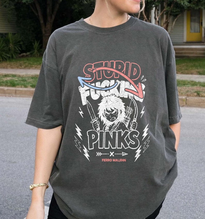 May include: Dark gray t-shirt with a graphic design. The design features the words "STUPID FUCKING PINKS" in red and white, with a cartoon character holding knives and arrows. The shirt also has lightning bolt graphics and the text "FERRO MALINN".