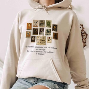 May include: Beige hoodie with a collection of vintage-style postage stamp graphics. The stamps feature various illustrations and the text "BE WARY DE CLEVER BE GOOD". Below the stamps is a quote about wind, trees, shadows, and nightmares.
