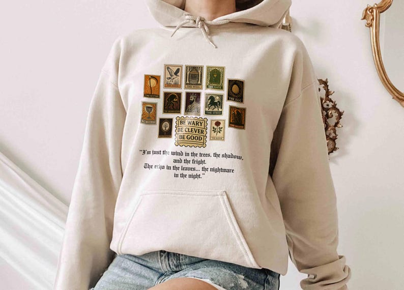 May include: Beige hoodie with a collection of vintage-style postage stamp graphics. The stamps feature various illustrations and the text "BE WARY DE CLEVER BE GOOD". Below the stamps is a quote about wind, trees, shadows, and nightmares.