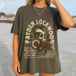 May include: Dark gray t-shirt with a graphic design featuring a skull, snake, compass, and red flower. The text "KAYDEN LOCKWOOD" arches above the design, with additional text below. A casual, vintage-style tee.