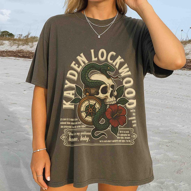 May include: Dark gray t-shirt with a graphic design featuring a skull, snake, compass, and red flower. The text "KAYDEN LOCKWOOD" arches above the design, with additional text below. A casual, vintage-style tee.