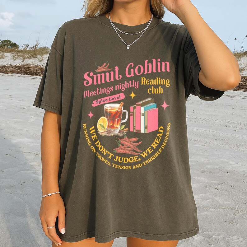 May include: Dark olive green t-shirt with a graphic that reads "Smut Goblin Meetings nightly Reading club". The graphic includes a cocktail, books, and chili peppers. The text "We don't judge, we read" is also included.